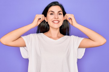 Canvas Print - Young beautiful brunette woman wearing casual white t-shirt over purple background Smiling pulling ears with fingers, funny gesture. Audition problem