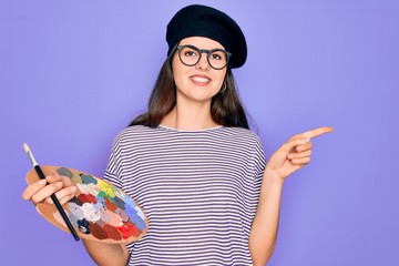 Canvas Print - Young beautiful artist girl wearing fashion beret painting using paintbrush and palette very happy pointing with hand and finger to the side