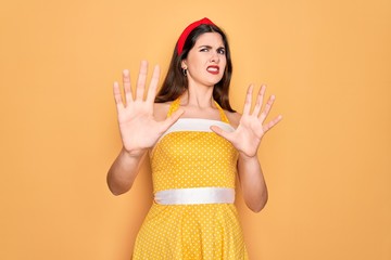 Canvas Print - Young beautiful pin up woman wearing 50s fashion vintage dress over yellow background afraid and terrified with fear expression stop gesture with hands, shouting in shock. Panic concept.