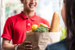 Asian deliver man worker in red color uniform handling bag of food, fruit, vegetable give to young beautiful female costumer in front of the house. Postman and express grocery delivery service concept