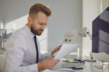 Wall Mural - Serious busy businessman reads working documents sitting at a table with a computer in the office.