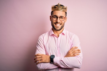 Young handsome business man wearing golden crown as king over pink background happy face smiling with crossed arms looking at the camera. Positive person.