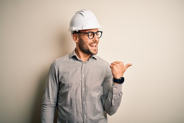 Canvas Print - Young architect man wearing builder safety helmet over isolated background smiling with happy face looking and pointing to the side with thumb up.