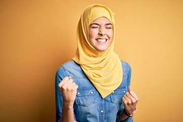 Canvas Print - Young beautiful girl wearing muslim hijab standing over isolated yellow background very happy and excited doing winner gesture with arms raised, smiling and screaming for success. Celebration concept.