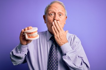 Poster - Grey haired senior man holding orthodontic prosthesis denture over purple background cover mouth with hand shocked with shame for mistake, expression of fear, scared in silence, secret concept