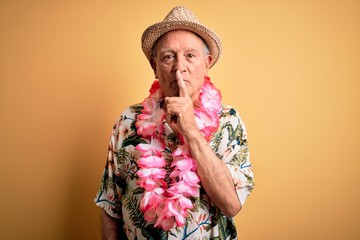 Canvas Print - Grey haired senior man wearing summer hat and hawaiian lei over yellow background asking to be quiet with finger on lips. Silence and secret concept.