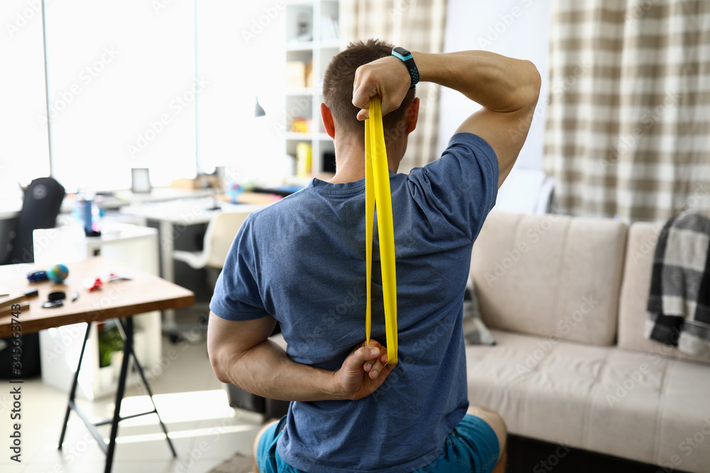 Close-up of man doing exercise for back using fitness rubber band. Home workout on quarantine period. Cozy living room interior. Strong and fit body. Sport and active lifestyle concept