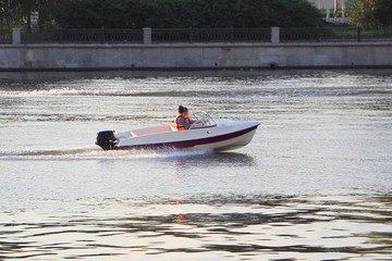 Wall Mural - One non-registrated 9.9 hp plastic outboard motor boat with two passengers in life jackets floats on the river against the background of the city embankment on a summer day