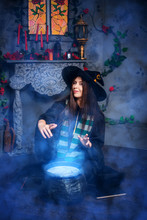Halloween Sorceress Concept. A Witch In A Black Robe And Hat Conjures Her Hands Over A Pot Of Boiling Potion, Sitting In Blue Magic Smoke.