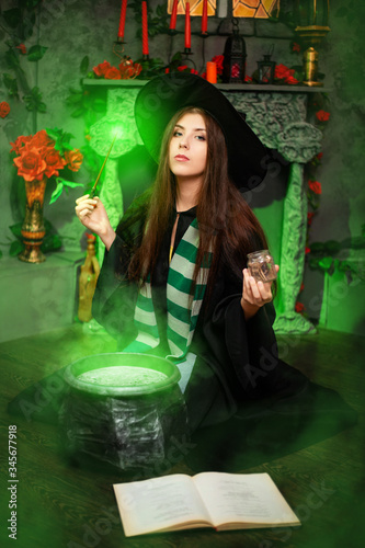 Fotografía A young witch in a black hat and mantle makes a potion in a black cauldron with a magic wand and a book