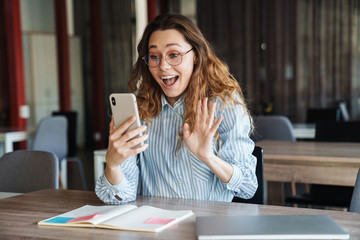 Canvas Print - Image of joyful woman waving hand and using cellphone while studying