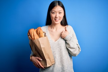 Sticker - Young asian woman holding bread grocery paper bag over blue isolated background happy with big smile doing ok sign, thumb up with fingers, excellent sign