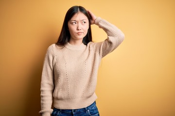 Wall Mural - Young beautiful asian woman wearing casual sweater over yellow isolated background confuse and wondering about question. Uncertain with doubt, thinking with hand on head. Pensive concept.