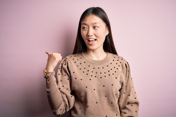 Wall Mural - Young beautiful asian woman wearing fashion and elegant sweater over pink solated background smiling with happy face looking and pointing to the side with thumb up.