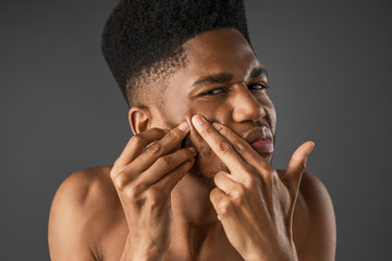 African man squeezes acne on face isolated on dark grey background. Skin care concept