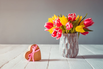 Bouquet of pink and yellow tulip flowers in a blue vase and heart shape gift box with satin ribbon bow on the white wooden table and grey background. Greetings for mother's or women's day. Card mockup