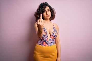 Wall Mural - Young beautiful arab woman on vacation wearing swimsuit and sunglasses over pink background Showing middle finger, impolite and rude fuck off expression