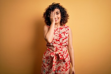 Sticker - Young beautiful curly arab woman on vacation wearing summer floral dress and sunglasses covering one eye with hand, confident smile on face and surprise emotion.