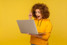 Wondered Excited Bright Woman With Curly Hair In Urban Style Hoody Holding Laptop And Pointing Finger Up, Inspired By Great Idea, Smart Solution. Indoor Studio Shot Isolated On Yellow Background