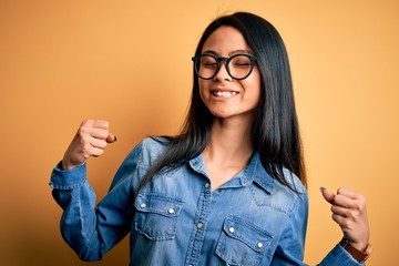 Wall Mural - Young beautiful chinese woman wearing casual denim shirt over isolated yellow background very happy and excited doing winner gesture with arms raised, smiling and screaming for success. Celebration