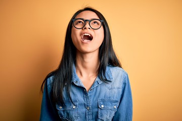 Wall Mural - Young beautiful chinese woman wearing casual denim shirt over isolated yellow background angry and mad screaming frustrated and furious, shouting with anger. Rage and aggressive concept.