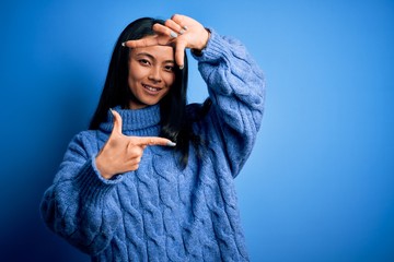 Wall Mural - Young beautiful chinese woman wearing casual sweater over isolated blue background smiling making frame with hands and fingers with happy face. Creativity and photography concept.