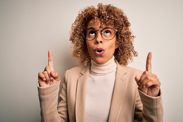 Beautiful african american businesswoman wearing glasses over isolated white background amazed and surprised looking up and pointing with fingers and raised arms.