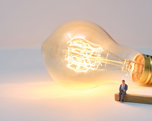 A businessman sits on a wooden bench while working on a laptop with a large lit filament lightbulb behind him - Tiny People Working From Home 