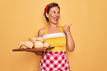 Poster - Middle age senior pin up housewife woman wearing 50s style retro dress and apron cooking bread pointing and showing with thumb up to the side with happy face smiling