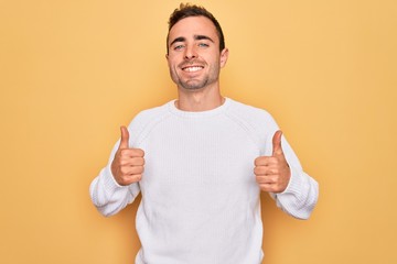 Young handsome man with blue eyes wearing casual sweater standing over yellow background success sign doing positive gesture with hand, thumbs up smiling and happy. Cheerful expression and winner