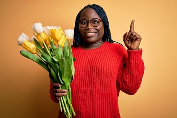 Wall Mural - Young african american plus size woman with braids holding bouquet of yellow tulips flower pointing finger up with successful idea. Exited and happy. Number one.