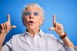 © Krakenimages.com - Senior beautiful woman wearing elegant shirt and glasses over isolated blue background amazed and surprised looking up and pointing with fingers and raised arms.