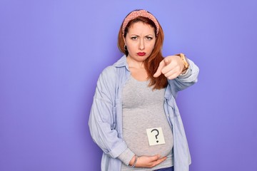 Poster - Young beautiful redhead pregnant woman expecting baby with question mark on stomach pointing with finger to the camera and to you, hand sign, positive and confident gesture from the front