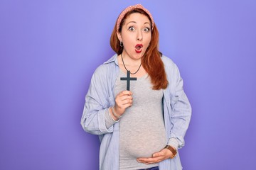 Wall Mural - Young beautiful redhead pregnant woman expecting baby holding christian cross scared in shock with a surprise face, afraid and excited with fear expression