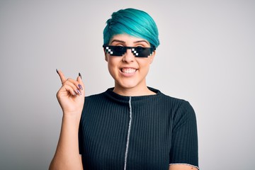 Sticker - Young woman with blue fashion hair wearing thug life sunglasses over white background with a big smile on face, pointing with hand and finger to the side looking at the camera.