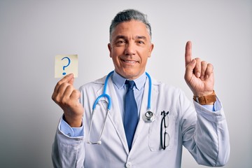 Sticker - Middle age handsome grey-haired doctor man holding paper with question mark surprised with an idea or question pointing finger with happy face, number one