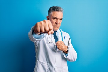 Poster - Middle age handsome grey-haired doctor man wearing coat and blue stethoscope Punching fist to fight, aggressive and angry attack, threat and violence