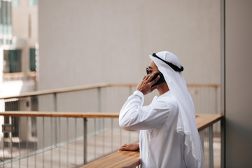 Wall Mural - Emirati guy wearing traditional kandura in urban city emirates lifestyle talking to smart phone. Side view photography concept for Arab magazine.