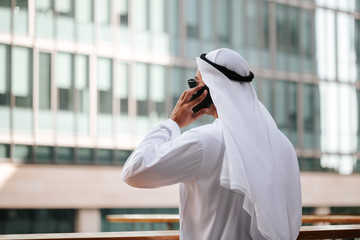 Wall Mural - Emirati guy wearing traditional kandura in urban city emirates lifestyle talking to smart phone. Overlooking building from the balcony. Photography concept for Arab magazine.