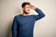 Young Handsome Man With Beard Wearing Casual Sweater Standing Over White Background Very Happy And Smiling Looking Far Away With Hand Over Head. Searching Concept.