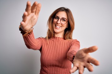 Young beautiful brunette woman wearing casual sweater and glasses over white background looking at the camera smiling with open arms for hug. Cheerful expression embracing happiness.