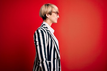 Poster - Blonde business woman with short hair wearing glasses and striped jacket over red background looking to side, relax profile pose with natural face with confident smile.