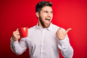 Young blond businessman with beard and blue eyes drinking red cup of coffee pointing and showing with thumb up to the side with happy face smiling