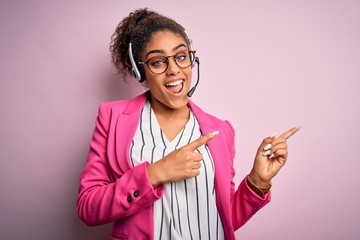 Canvas Print - Young african american call center agent girl wearing glasses working using headset smiling and looking at the camera pointing with two hands and fingers to the side.