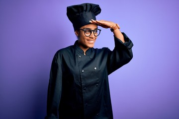 Sticker - Young african american chef girl wearing cooker uniform and hat over purple background very happy and smiling looking far away with hand over head. Searching concept.