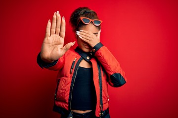 Sticker - Beautiful african american girl wearing red jacket and sunglasses over isolated background covering eyes with hands and doing stop gesture with sad and fear expression. Embarrassed and negative