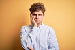 © Krakenimages.com - Young blond handsome man with curly hair wearing striped shirt over yellow background thinking looking tired and bored with depression problems with crossed arms.