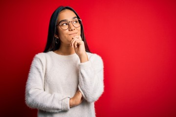 Wall Mural - Young beautiful asian woman wearing casual sweater and glasses over red background with hand on chin thinking about question, pensive expression. Smiling and thoughtful face. Doubt concept.
