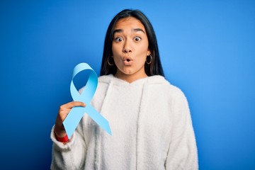 Wall Mural - Young asian woman holding blue cancer ribbon symbol standing over isolated background scared in shock with a surprise face, afraid and excited with fear expression