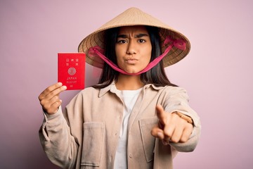 Canvas Print - Young thai woman wearing traditional conical asian hat holding japan japanese passport pointing with finger to the camera and to you, hand sign, positive and confident gesture from the front